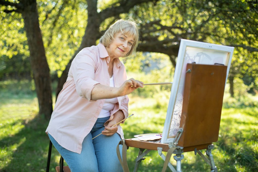 happy caucasian retired woman wearing light shirt and denim jeans painting on canvas and having fun at beautiful green garden or park on sunny day art hobby and retirement